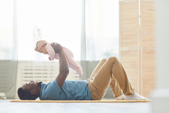 Young African father lying on the floor and exercising together with his baby girl at home