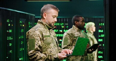 Caucasian military male officer in camouflage standing in server room with laptop computer and checking data on monitors. Work with secret info in army. Digital concept. African American man behind.