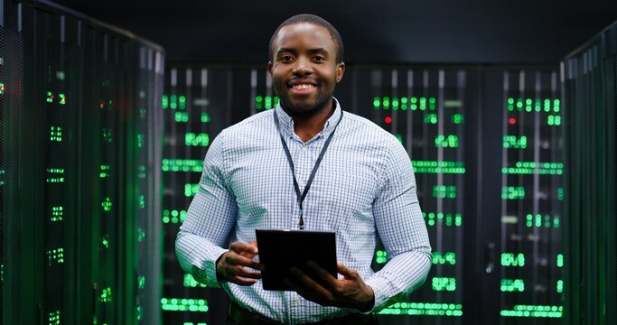 Portrait of young handsome African American man smiling cheerfully too camera in database center full of computers and servers. Close up of male worker of server room. Cybersecurity world.