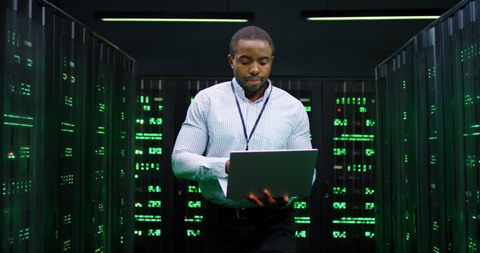 Male African American big data analytic walking at server dark room, working with secret data and using laptop computer to check the information. Monitoring database center. Cloud storage of info.