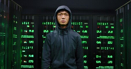Portrait of Asian young hacker in hood walking among severs in dark room and looking at camera. Guy...