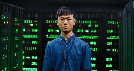 Portrait of young Asian guy looking at camera with slight smile and standing among servers in dark room. Male cybesecurity developer posing among computers.