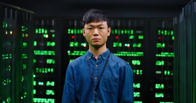 Portrait of young Asian guy looking at camera with slight smile and standing among servers in dark room. Male cybesecurity developer posing among computers.