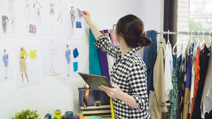 Young fashion designer asian japanese woman looking at her tablet thinking details creating in studio. dressmaker hold digital touchpad choosing color of fabric textile cloth sample on wall sketch