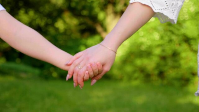 Close up mom and son hold hands, walk along the grass in a botanical garden. Woman and a boy are walking in the forest against a background of green trees. 4K slow motion footage
