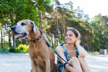 Beautiful young girl alone with her dog sitting on the beach. Cute smiling woman and Rhodesian Ridgeback pet are best friends.
