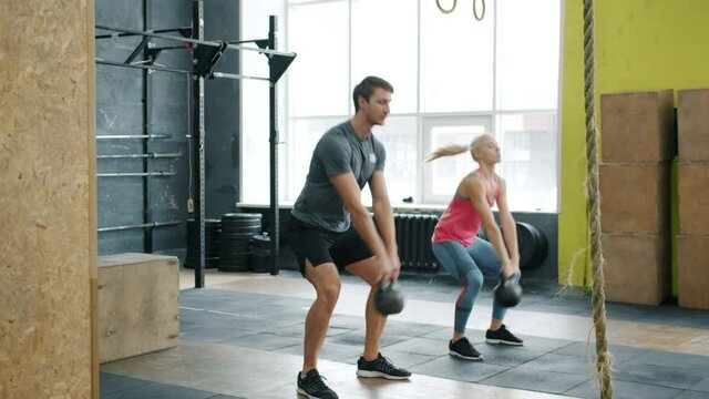 Slow motion of young couple guy and girl exercising in gym squatting with kettlebells working out together. Crossfit, active lifestyle and people concept.