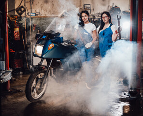 Obraz premium Two young female mechanic posing next to a sportbike in authentic workshop garage surrounded by smoke