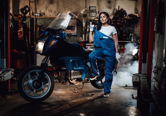 Naklejka na ściany i meble Beautiful brunette woman in blue overalls posing with big wrench while standing next to a sportbike in garage or workshop