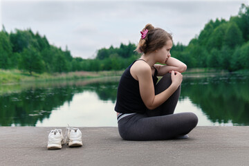 Teenage girl in a bad mood sitting on pond pier in cloudy summer morning. The girl is worried about something. Awkward age.