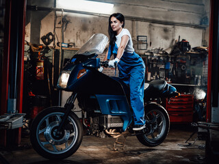 Obraz na płótnie Canvas Young beautiful female mechanic in blue overalls posing on sportbike in garage or workshop