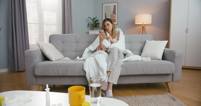Unhealthy young Caucasian woman with fever sitting on couch in living room and videochatting on smartphone. Sick girl caughing and having videochat on mobile phone via webcam.