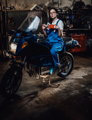 Obraz na płótnie Canvas Beautiful young brunette female mechanic in blue overalls sitting on sportbike in garage or workshop