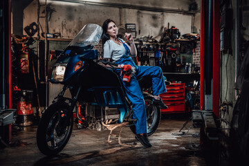 Plakat Hot brunette woman in blue overalls posing for a camera while leanign on sportbike in garage or workshop