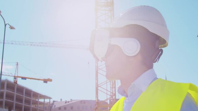 Professional builder in VR helmet standing in front of construction site and using virtual and augmented reality technologies. Office building and crane background. Real estate and investment.