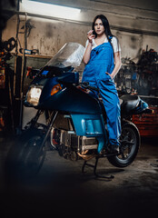 Young female mechanic relaxing smoking a cigarette while standing on sportbike in garage or workshop