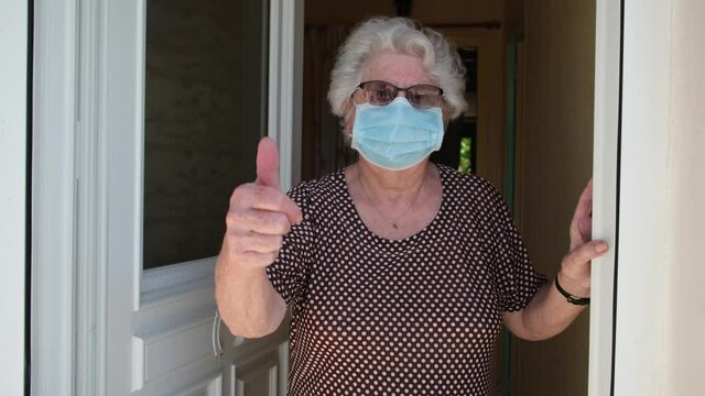 elderly old senior woman opening front door of house and welcoming people at home wearing a surgical mask to avoid covid19 and coronavirus contamination