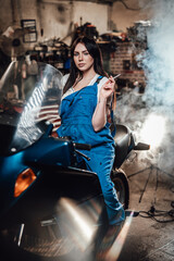 Fototapeta na wymiar Beautiful brunette female mechanic in blue overalls relaxing smoking a cigarette while sitting on sportbike in garage or workshop