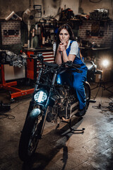 Beautiful brunette female mechanic in blue overalls relaxing smoking a cigarette while sitting on custom bobber in garage or workshop