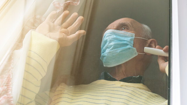 pensioner in blue mask stands at window waving hand