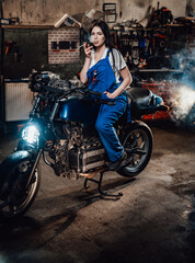 Fototapeta na wymiar Beautiful brunette female mechanic in blue overalls relaxing smoking a cigarette while sitting on custom bobber in garage or workshop