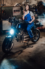 Plakat Beautiful brunette female mechanic in blue overalls relaxing smoking a cigarette while sitting on custom bobber in garage or workshop