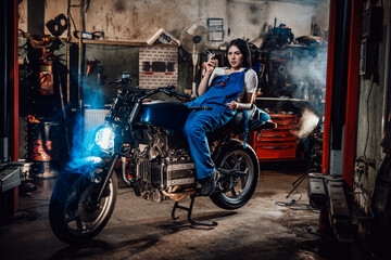Fototapeta na wymiar Hot brunette female mechanic in blue overalls relaxing smoking a cigarette while lying on custom bobber in garage or workshop