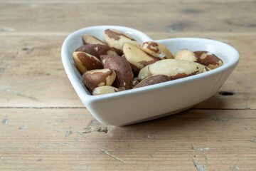 A bowl of Brazil Nuts in a white heart shaped ceramic bowl on an oak tabletop