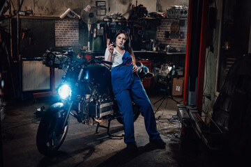 Fototapeta na wymiar Woman mechanic relaxing smoking a cigarette while leaning on custom bobber in garage or workshop