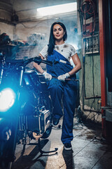 Naklejka na ściany i meble Beautiful brunette woman in blue overalls posing with big wrench while standing next to a custom bobber in garage or workshop