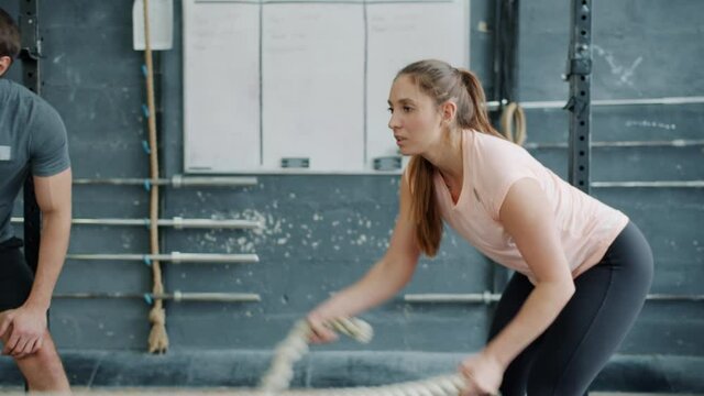 Fit girl in sportswear is training with battle ropes in gym while male trainer is assisting motivating speaking and gesturing. Millennials and sports concept.
