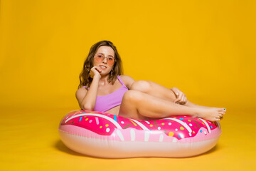 Obraz na płótnie Canvas Positive millennial blonde sitting on big inflatable ring, ready to swim, yellow studio background