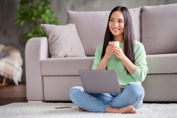 Full size photo of positive asian girl work remote laptop dream dreamy weekend hold mug beverage sit floor carpet legs crossed folded in house indoors