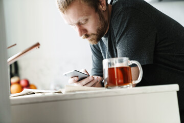 Photo of serious redhead man using mobile phone while drinking tea