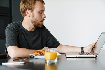 Photo of caucasian young man using laptop while having breakfast