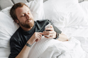 Photo of pleased bearded man using mobile phone while lying after sleep