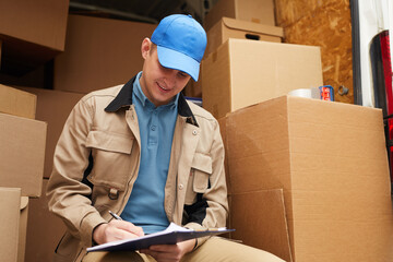 Delivery person filling the documents while sitting outdoors with cardboard boxes in the background