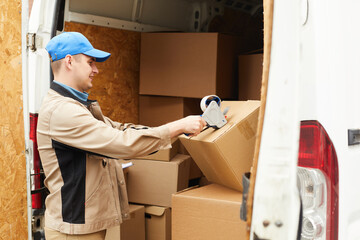 Young man in uniform packing the cardboard boxes with adhesive tape before shipping