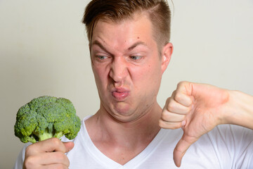 Portrait of young handsome man holding broccoli and giving thumbs down