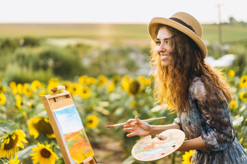 A young woman with curly hair and wearing a hat is painting in nature. A woman stands in a sunflower field on a beautiful day