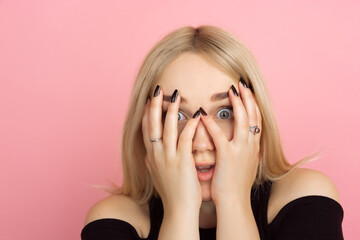 Shocked hiding face. Portrait of young caucasian woman with bright emotions on coral pink studio background. Blonde female model. Concept of human emotions, facial expression, sales, advertising