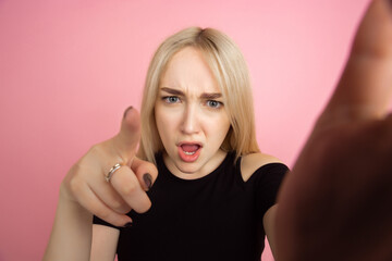 Selfie view. Portrait of young caucasian woman with bright emotions on coral pink studio background. Blonde female model. Concept of human emotions, facial expression, sales, advertising, youth.