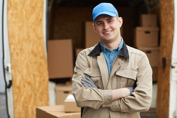 Portrait of young delivery person in uniform standing with arms crossed and smiling at camera while working outdoors