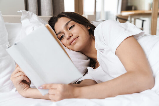 Photo of pretty smiling woman reading book while lying in bed