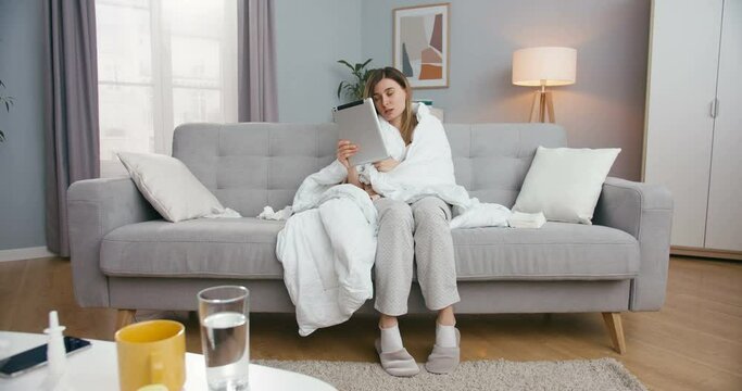 Unhealthy young Caucasian woman with fever sitting on couch in living room and videochatting on tablet. Sick girl caughing and having videochat on computer via webcam Female talking in video on device