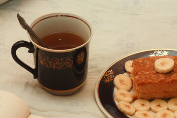 A blue cup of hot tea, a piece of shortbread cake with condensed milk and bananas, cut rings on a saucer and an open book with white blank pages on sheets on the bed.