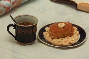 A blue cup of hot tea, a piece of shortbread cake with condensed milk and bananas, cut rings on a saucer and an open book with white blank pages on sheets on the bed.