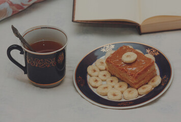 A blue cup of hot tea, a piece of shortbread cake with condensed milk and bananas, cut rings on a saucer and an open book with white blank pages on sheets on the bed.