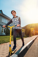 A teenage boy with a serious face rides a scooter on the sidewalk. In the background, an empty street. Bottom view. Concept of sports recreation and youth activity