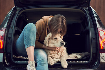 Young woman hugs golden retriever while sitting in open trunk of black car